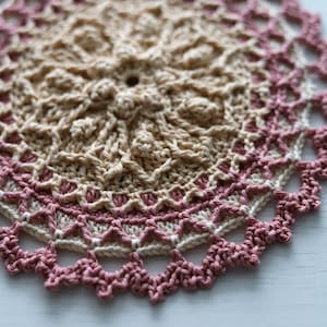 K&ouml;nnte beinhalten: Eine geh&auml;kelte Tischdecke mit einem beigefarbenen Zentrum und einem rosa Rand. Die Tischdecke ist mit einem filigranen Stichmuster gearbeitet und hat einen gewellten Rand.