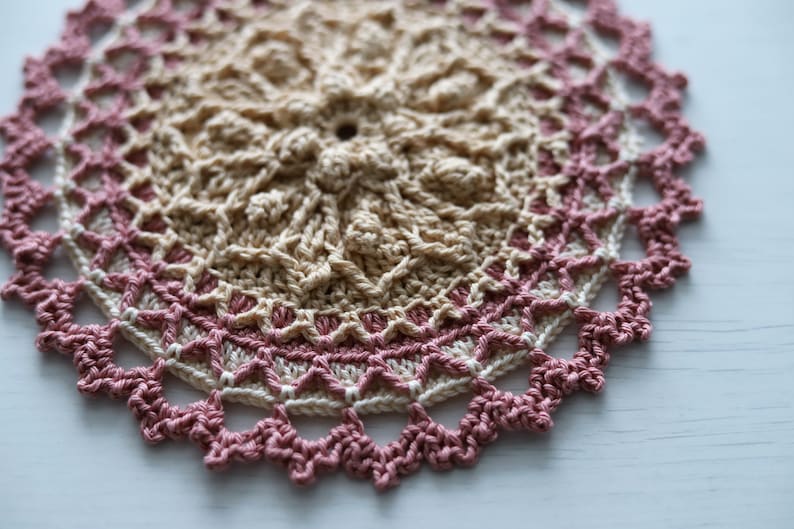 K&ouml;nnte beinhalten: Eine geh&auml;kelte Tischdecke mit einem beigefarbenen Zentrum und einem rosa Rand. Die Tischdecke ist mit einem filigranen Stichmuster gearbeitet und hat einen gewellten Rand.