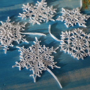 May include: Five white crocheted snowflakes on a blue background. The snowflakes are delicate and intricate, with a variety of patterns.