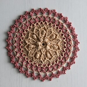 K&ouml;nnte beinhalten: Eine geh&auml;kelte Tischdecke mit einem runden Design in Beige- und Rosat&ouml;nen. Die Tischdecke hat einen zarten Spitzenrand.