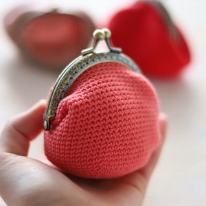 May include: A small, crocheted coin purse with a metal clasp. The purse is a bright coral color and is held in a hand.