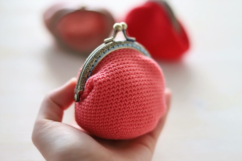 May include: A small, crocheted coin purse with a metal clasp. The purse is a bright coral color and is held in a hand.