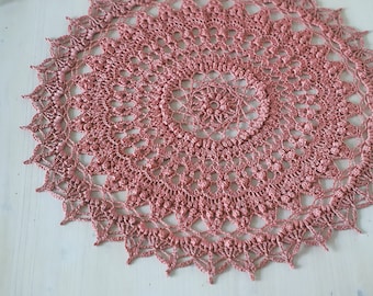 Puede incluir: Un delicado encaje de ganchillo en color rosa polvoriento. El encaje es circular y presenta un diseño intrincado y de encaje.