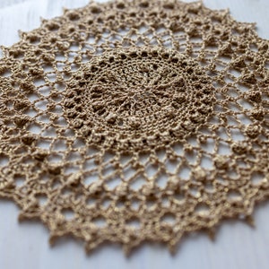 May include: A delicate, crocheted doily in a light beige color. The doily has a circular shape with a intricate pattern of woven stitches.