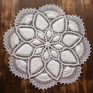 May include: White crocheted doily with a delicate floral design. The doily has a circular shape and is made with intricate stitches.