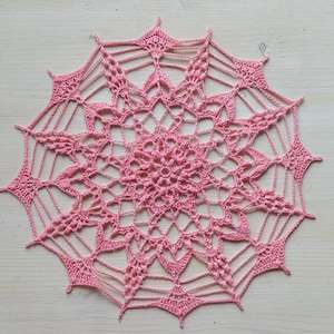 May include: A pink crocheted doily with a delicate, intricate design. The doily has a starburst pattern in the center and a circular shape.