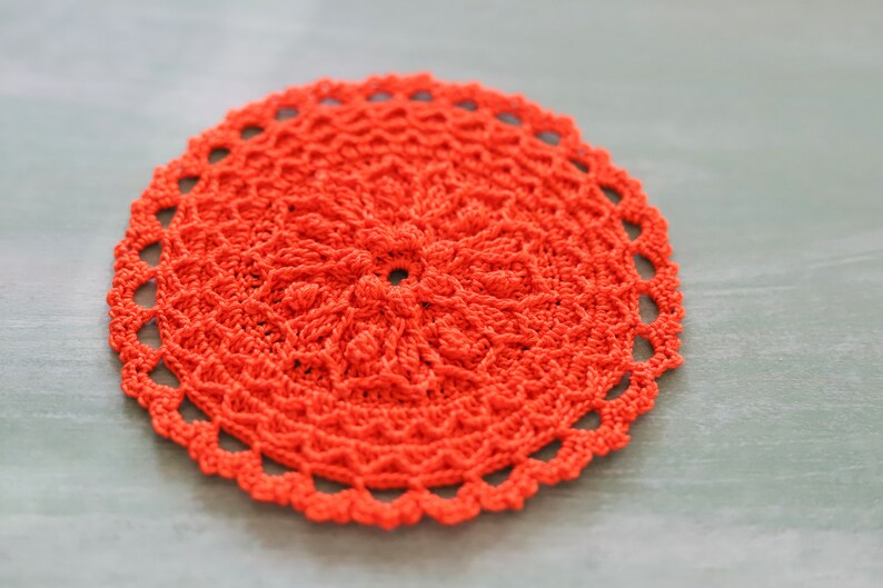 K&ouml;nnte beinhalten: Eine leuchtend orangefarbene geh&auml;kelte Tischdecke mit einem runden Design. Die Tischdecke hat eine zarte Spitzenborte am Rand.