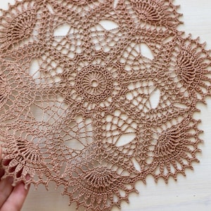Può includere: Un delicato tovagliolo all'uncinetto marrone con un design circolare che presenta intricati motivi floreali.