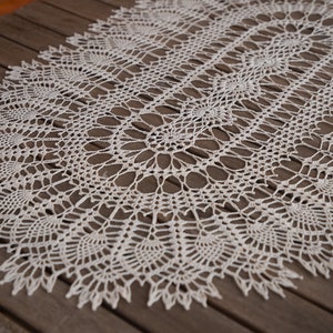 Può includere: Un runner da tavolo in crochet bianco con un delicato motivo floreale. Il runner è disposto su una superficie in legno.