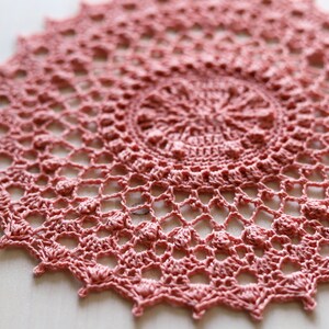 May include: A delicate pink crocheted doily with a circular design and scalloped edges.
