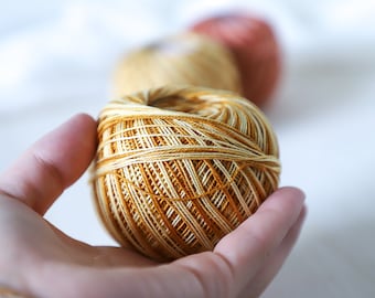 May include: A ball of yarn in shades of brown and cream, held in a hand. The yarn is wound tightly and has a textured appearance.