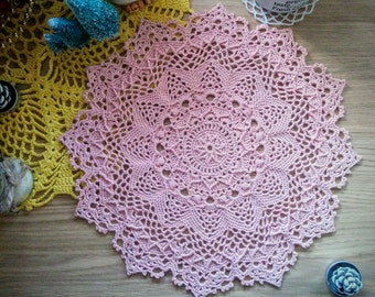 Puede incluir: Un delicado encaje de ganchillo rosa con un patrón floral detallado. El encaje es circular y tiene un borde festoneado.