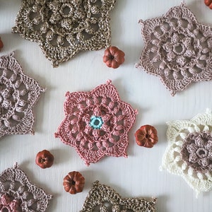 Op de afbeelding: Een verzameling van zes gehaakte stervormige doilies in verschillende tinten roze, bruin en wit. De doilies zijn gerangschikt op een wit oppervlak met kleine decoratieve pompoenen.
