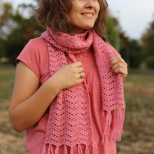 Op de afbeelding: Een roze gehaakte sjaal met franjes. De sjaal wordt gedragen door een persoon met bruin haar en een roze shirt.
