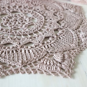 May include: A close-up of a crocheted doily in a light brown color. The doily has a delicate, intricate design with a circular center and a scalloped edge.