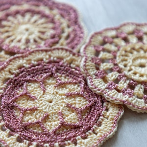 Può includere: Tre sottobicchieri all'uncinetto in uno schema di colori crema e rosa. I sottobicchieri hanno una forma circolare con un design centrale e un bordo smerlato.