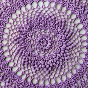 Può includere: Un primo piano di un centrino all'uncinetto viola con un design circolare. Il centrino ha un motivo delicato e intricato con un motivo floreale centrale.