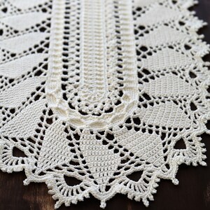 May include: A white crocheted table runner with a delicate lace pattern. The runner has a scalloped edge and is made with a fine yarn.