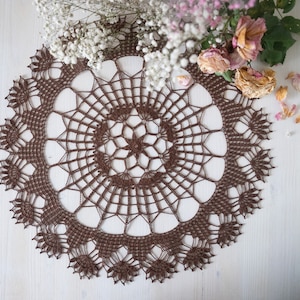 May include: A brown crocheted doily with a delicate floral pattern. The doily is circular and has a center with a star-shaped design.