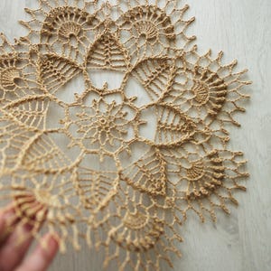 Può includere: Un centrino all'uncinetto beige fatto a mano. L'intricato design in pizzo presenta un motivo centrale a stella circondato da forme floreali e geometriche. Un delicato elemento di arredo per la casa.