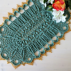Op de afbeelding: Een turquoise gehaakt tafellaken met een gouden gehaakte rand. Het tafellaken heeft een delicaat, ingewikkeld ontwerp met een gegolfde rand.