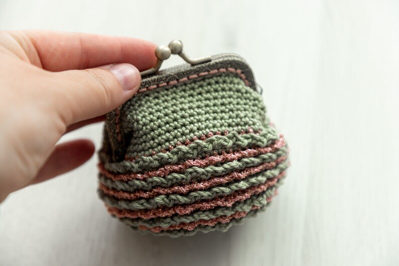 May include: A small crocheted coin purse with a green body and pink trim. The purse has a metal clasp.