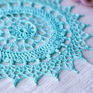 May include: A light blue crocheted doily with a delicate, intricate pattern. The doily is made with a fine yarn and has a circular shape.