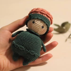 Puede incluir: Una muñeca de crochet con un vestido verde y un gorro rosa y verde. La muñeca tiene una cara blanca con ojos negros.