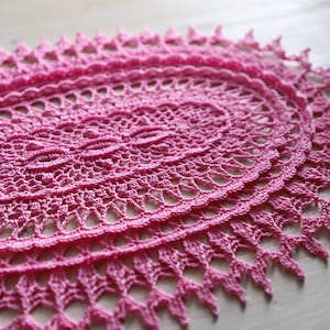 Op de afbeelding: Een roze gehaakte onderzetter met een delicaat bloemenpatroon. De onderzetter is ovaalvormig en heeft een gegolfde rand.