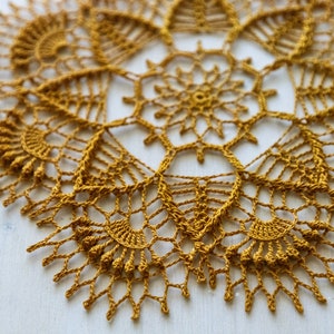 Può includere: Tovagliolo all'uncinetto giallo senape con un design traforato dettagliato. Il pezzo circolare presenta un motivo floreale centrale circondato da motivi intricati e un bordo smerlato. Il tessuto fatto a mano è esposto su una superficie bianca.