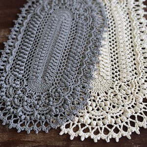 May include: A gray and white crocheted table runner with an oval design. The runner is made with a delicate stitch pattern and has a fringed edge.