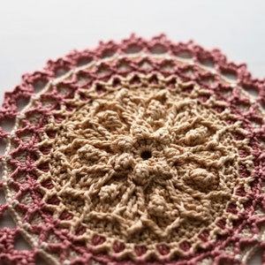 K&ouml;nnte beinhalten: Eine geh&auml;kelte Deckchen in Rosa und Beige. Das Deckchen ist rund und hat ein feines, detailliertes Muster.