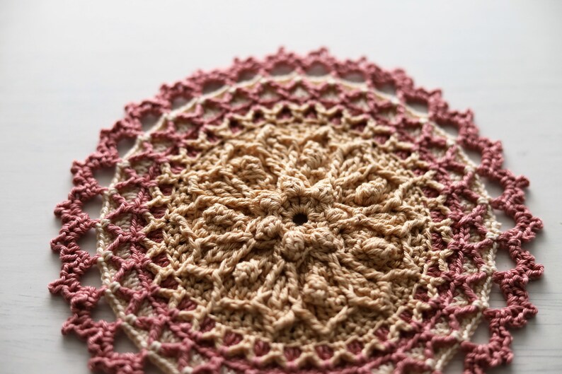 K&ouml;nnte beinhalten: Eine geh&auml;kelte Deckchen in Rosa und Beige. Das Deckchen ist rund und hat ein feines, detailliertes Muster.