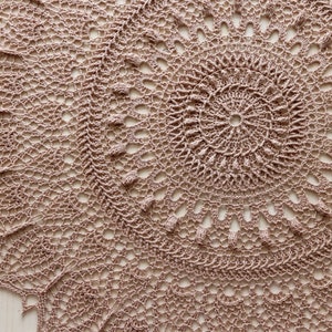 May include: A close-up of a beige crocheted doily with a circular design. The doily has a delicate, intricate pattern with multiple layers of crocheted stitches.