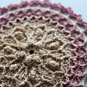 K&ouml;nnte beinhalten: Eine geh&auml;kelte Tischdecke mit einem beigefarbenen Zentrum und einem rosa-wei&szlig;en Rand.