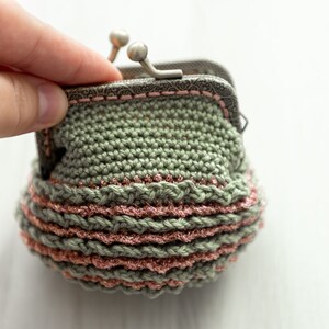 May include: A small crocheted coin purse with a green and pink striped design. The purse has a metal clasp and is open to reveal the inside.