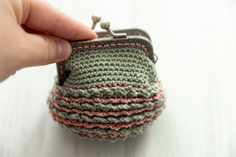 May include: A small crocheted coin purse with a green and pink striped design. The purse has a metal clasp and is open to reveal the inside.