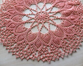 Puede incluir: Un delicado encaje de ganchillo de color rosa suave. El encaje tiene un diseño circular con un motivo floral central y un intrincado trabajo de encaje.