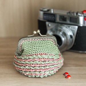 May include: A green crocheted coin purse with a pink trim and a silver clasp. The purse is sitting on a wooden surface next to a vintage camera.