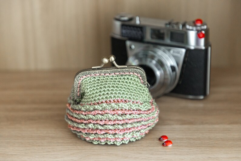 May include: A green crocheted coin purse with a pink trim and a silver clasp. The purse is sitting on a wooden surface next to a vintage camera.