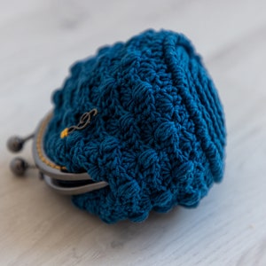 May include: A blue crocheted coin purse with a metal clasp. The purse is closed and lying on a white surface.