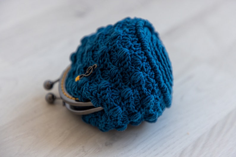 May include: A blue crocheted coin purse with a metal clasp. The purse is closed and lying on a white surface.