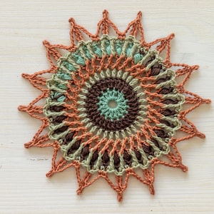 May include: A crocheted mandala with a brown, green, and orange color scheme. The mandala has a circular design with a central point and radiating spokes.