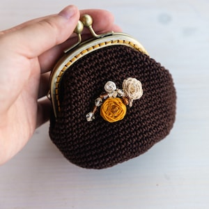 May include: A brown crocheted coin purse with a gold metal clasp and floral embellishments. The flowers are made of yellow, white, and brown yarn.