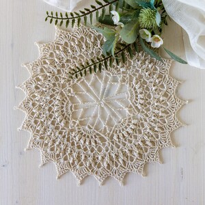 May include: A delicate, cream-colored crochet doily with a starburst pattern, laid on a white wooden surface.