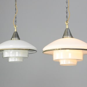 May include: Two white pendant lights with a tiered design and brass accents. The lights have a conical top and a circular shade. The lights are suspended by chains and are suitable for indoor lighting.