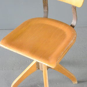 Op de afbeelding: Een vintage draaistoel met een houten zitting en een metalen frame. De stoel heeft een lichte houtkleur en een eenvoudig ontwerp.
