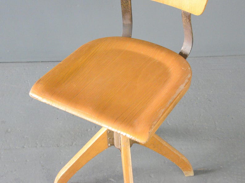 Op de afbeelding: Een vintage draaistoel met een houten zitting en een metalen frame. De stoel heeft een lichte houtkleur en een eenvoudig ontwerp.