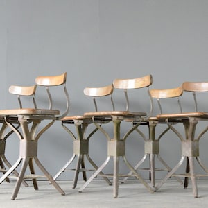 Puede incluir: Cinco sillas industriales vintage de metal y madera con estructura de metal gris y asiento de madera clara. Las sillas están dispuestas en fila y todas miran en la misma dirección.