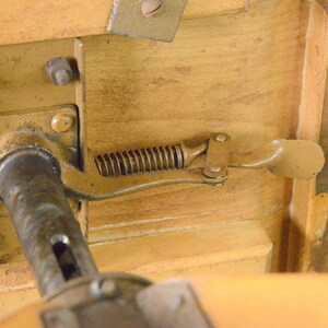 Op de afbeelding: Close-up van een houten tafel met een metalen veermechanisme. Het mechanisme heeft een metalen staaf en een hendel met een plat, rechthoekig pedaal. De tafel is gemaakt van licht hout en heeft een natuurlijke afwerking.